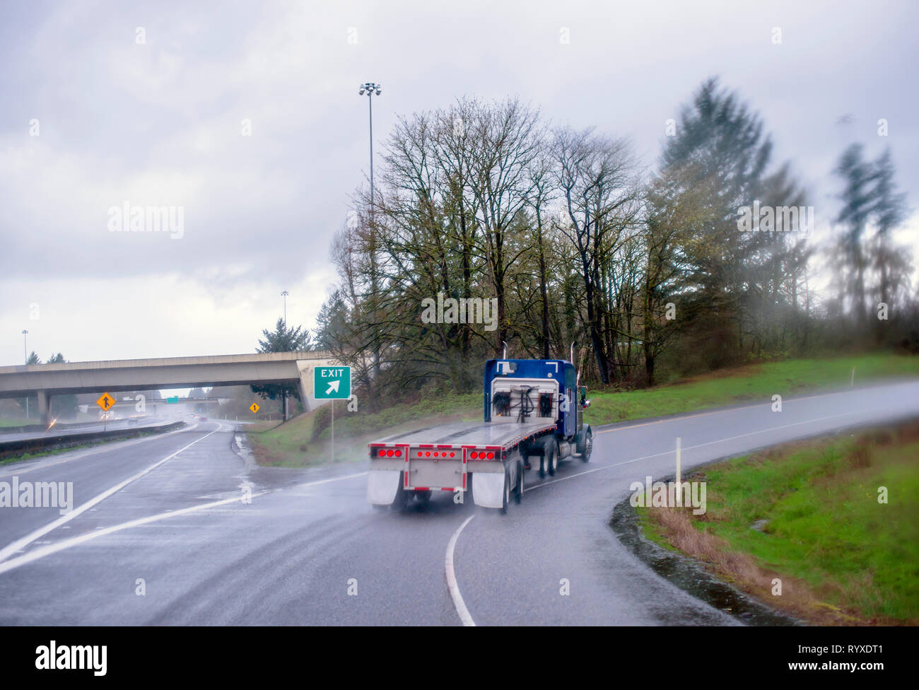 Big Rig blue American classic lungo il cofano motore trasportatore semi camion con pianale semi rimorchio spostando su autostrada uscita wet piove road con pioggia polvere sfocata h Foto Stock