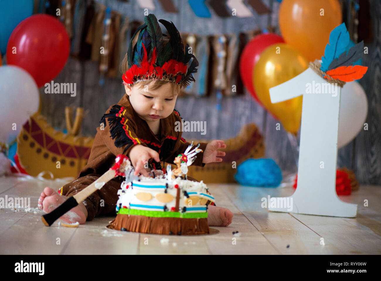 Foto riprese smash la torta per 1 anno in stile di capo indiano. Little Boy emotivo con un tomahawk in costume di Apache e in piuma copricapo Foto Stock