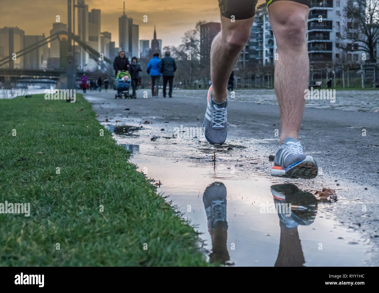 Vista delle gambe di un uomo che corre dopo la pioggia lungo un fiume in una grande città. Sport a molla Foto Stock