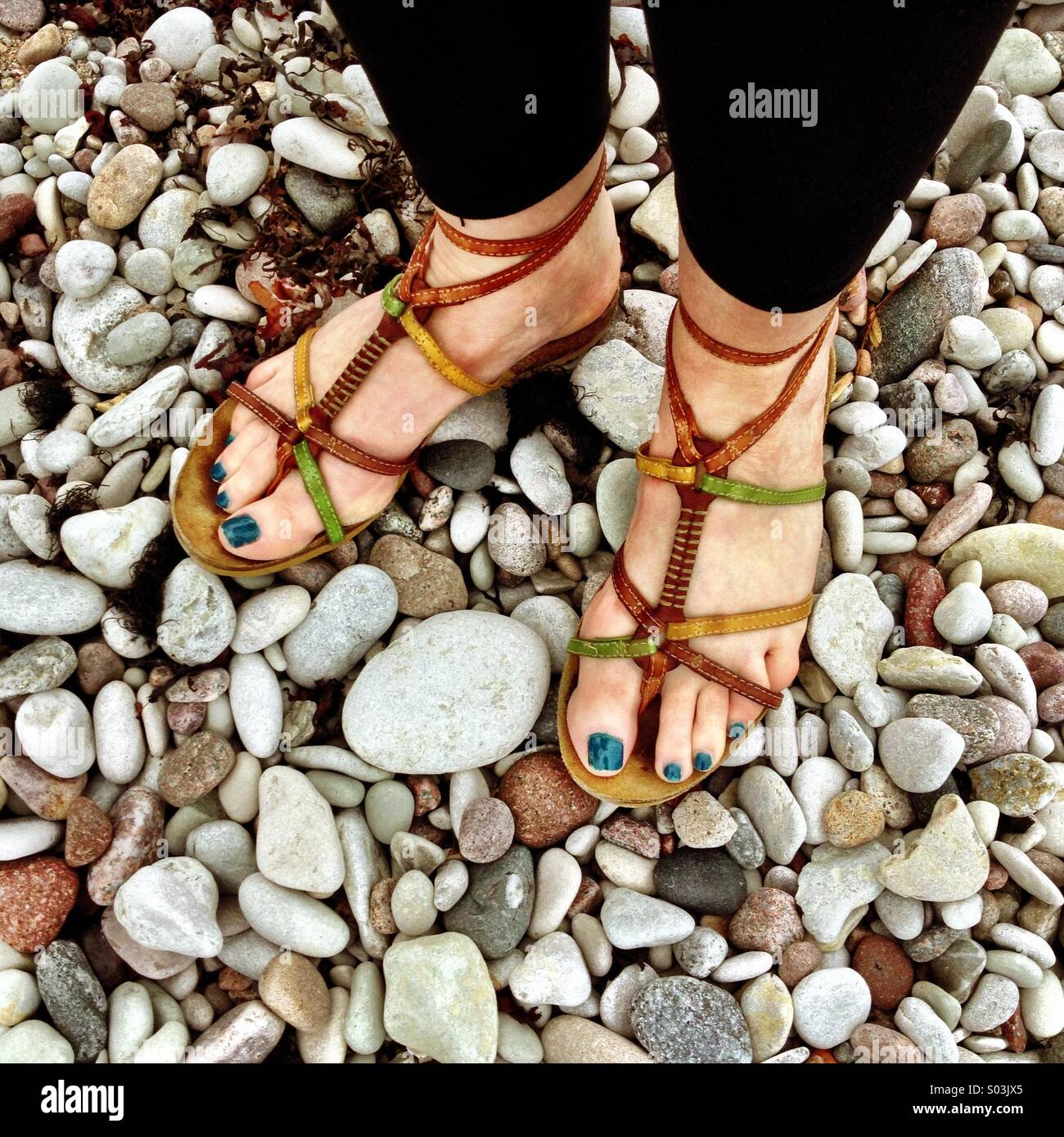 Piedi con blu unghie verniciate in sandali su di una spiaggia di ciottoli. Foto Stock