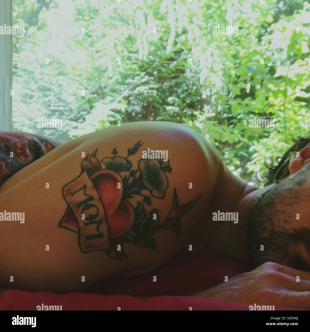 Scruffy uomo napping in letto nel pomeriggio con mom tattoo con finestra e alberi in background. Foto Stock