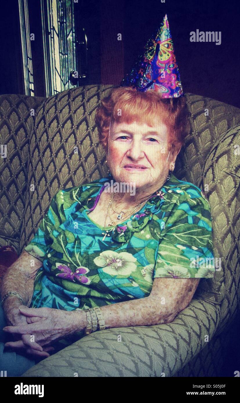Un senior citizen donna con i capelli rossi si siede su una sedia che indossa un cappello di compleanno Foto Stock