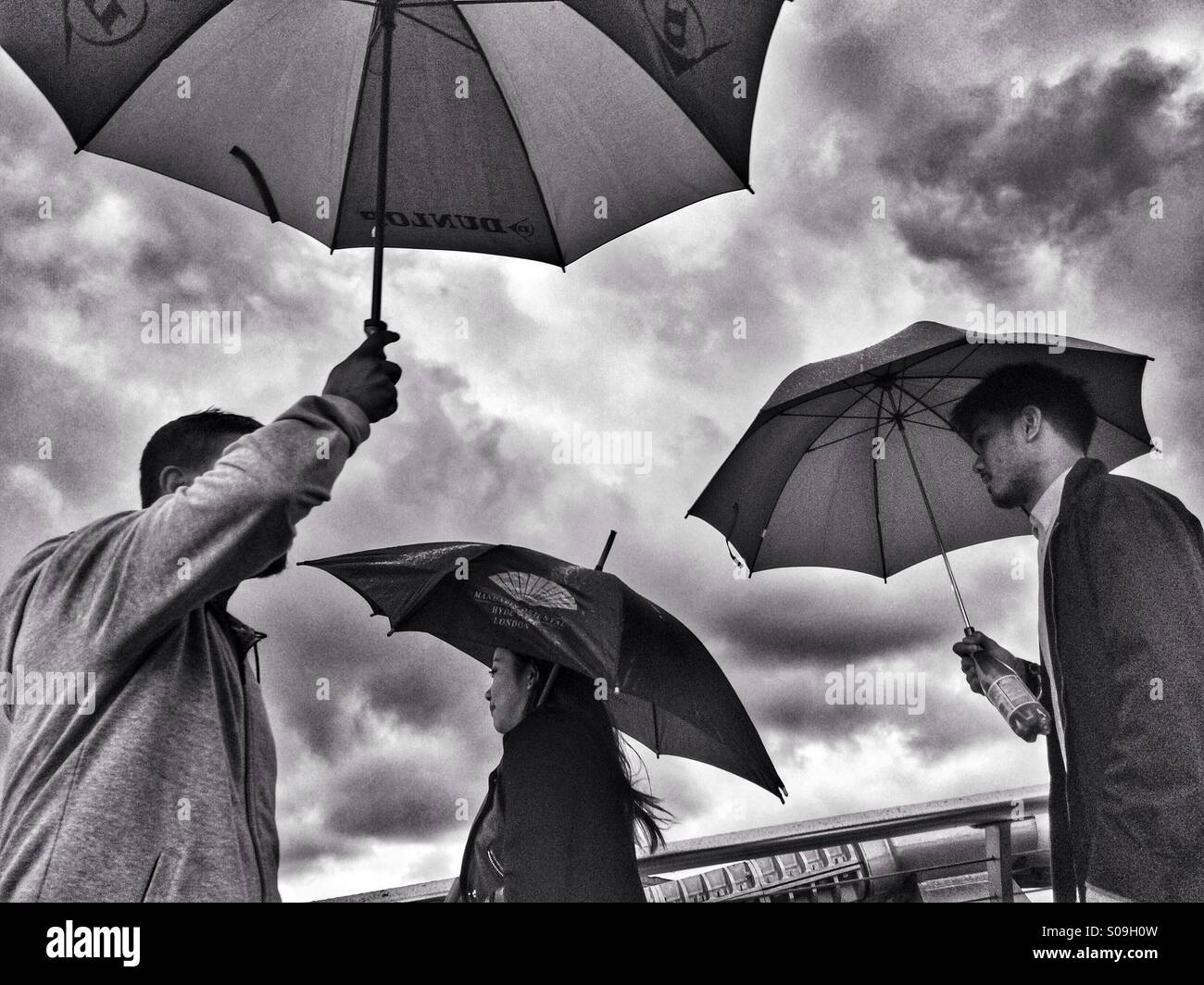 Foto in bianco e nero di tre persone ombrelli azienda tenta di camminare oltre ogni altro su un grigio e nuvoloso, giorno di pioggia a Londra. Foto Stock