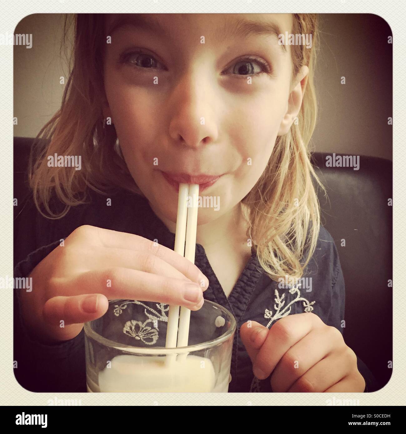 Un giovane biondo sorridente ragazza felicemente di bere da un bicchiere di latte usando una paglia Foto Stock