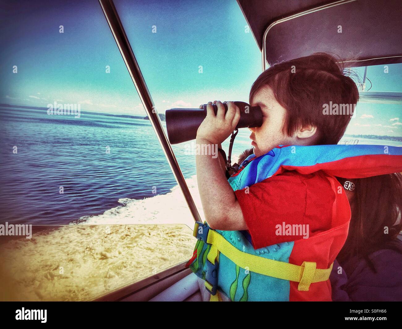 Un piccolo ragazzo su una barca di indossare un giubbotto di salvataggio si affaccia al mare con un paio di binocoli. Foto Stock