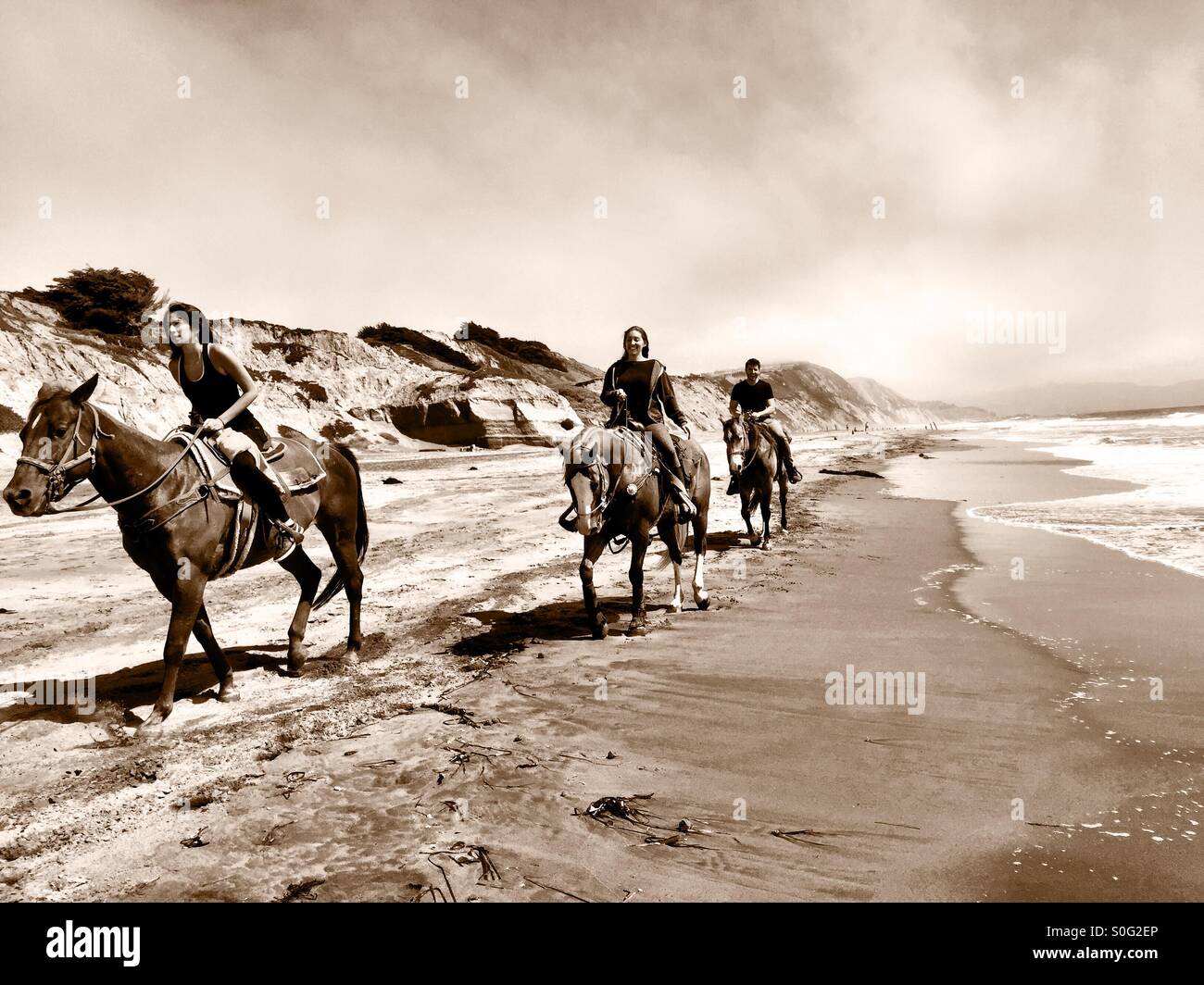 Tre i piloti a cavallo appaiono al di fuori di un lungo tratto di spiaggia della California, vanishing nel lontano foggy pacifico strato marino. Tonalità seppia. Foto Stock