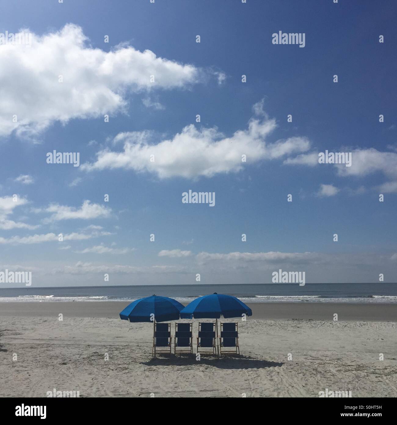 Per blue beach sedie e due ombrelli blu sedersi su un vuoto tratto di spiaggia sotto un luminoso cielo blu. Foto Stock