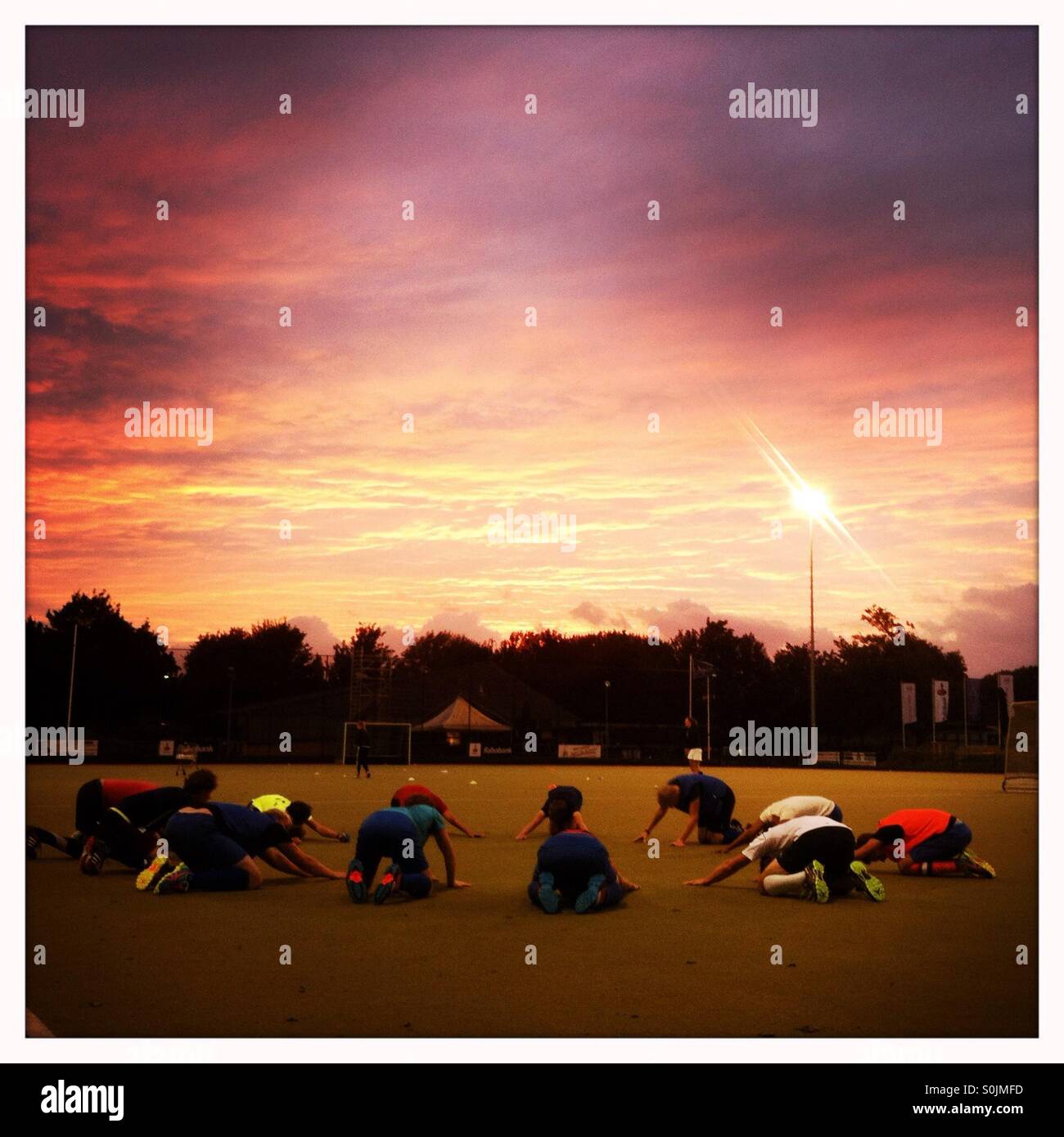 Una squadra di hockey facendo uno sport riscaldamento esercizio insieme in un cerchio con una drammatica rosso e viola il tramonto e il cielo dietro di loro. Foto Stock