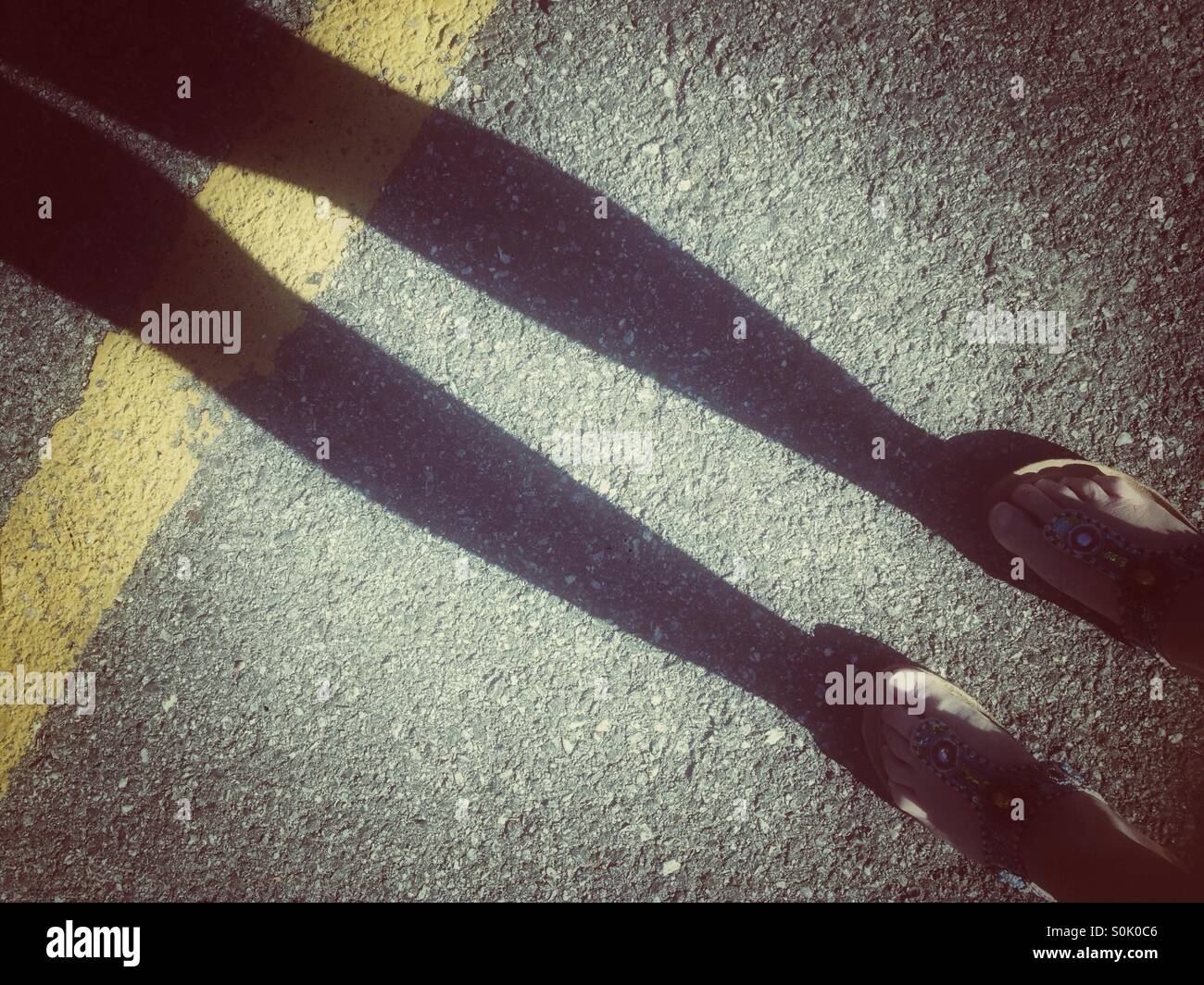 Ombra di donna di gambe e piedi in sandali estivi sulla strada con striscia gialla. Grunge retrò filtrato. Foto Stock