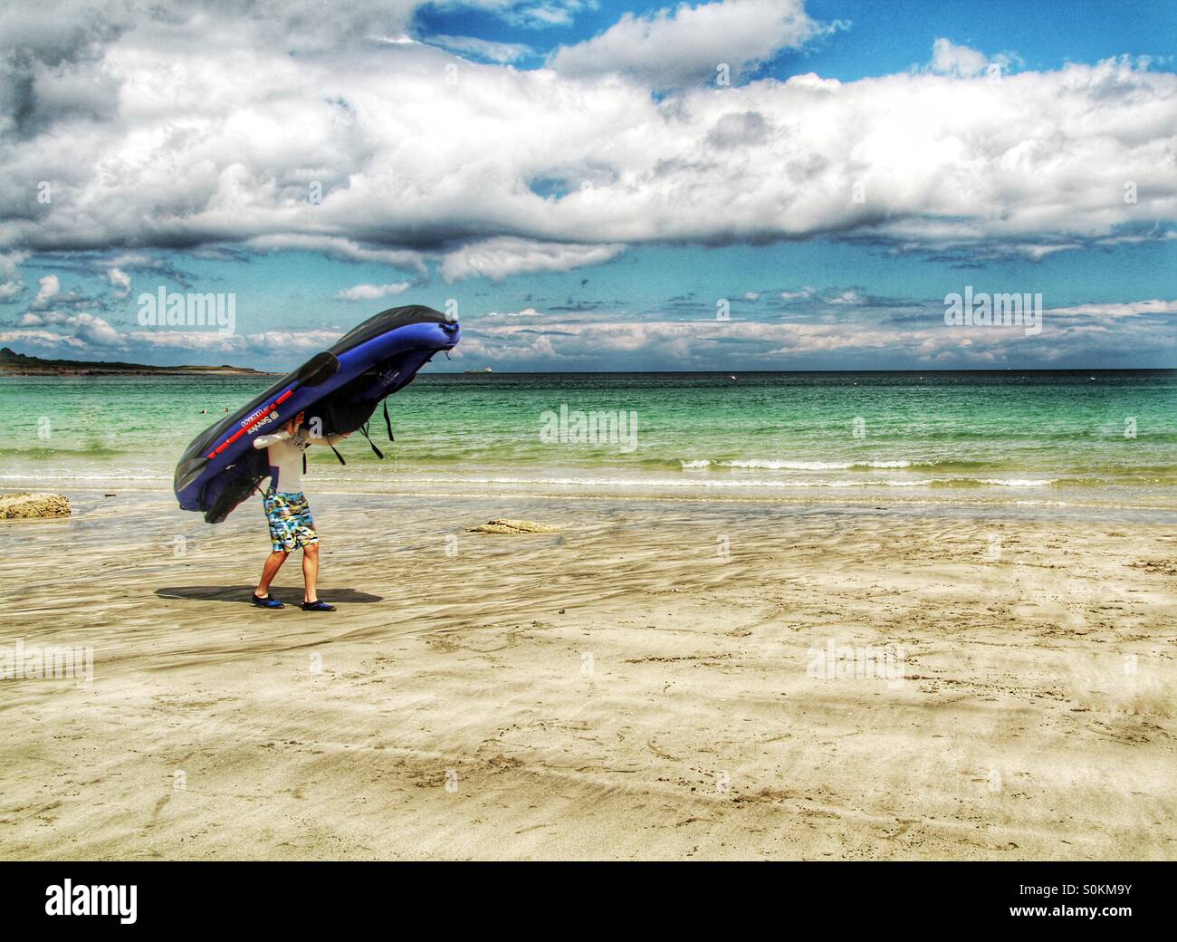 Un ragazzo che trasporta un gommone sulla sua testa come egli sta camminando lungo una spiaggia di sabbia in un giorno d'estate. Foto Stock