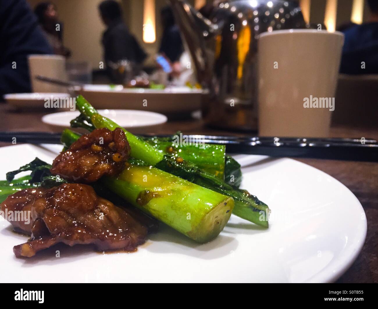 Vista ravvicinata del manzo e del gai-lan (broccoli cinesi) in un ristorante cinese su un piatto bianco, mentre altri commensali sono fuori fuoco Foto Stock