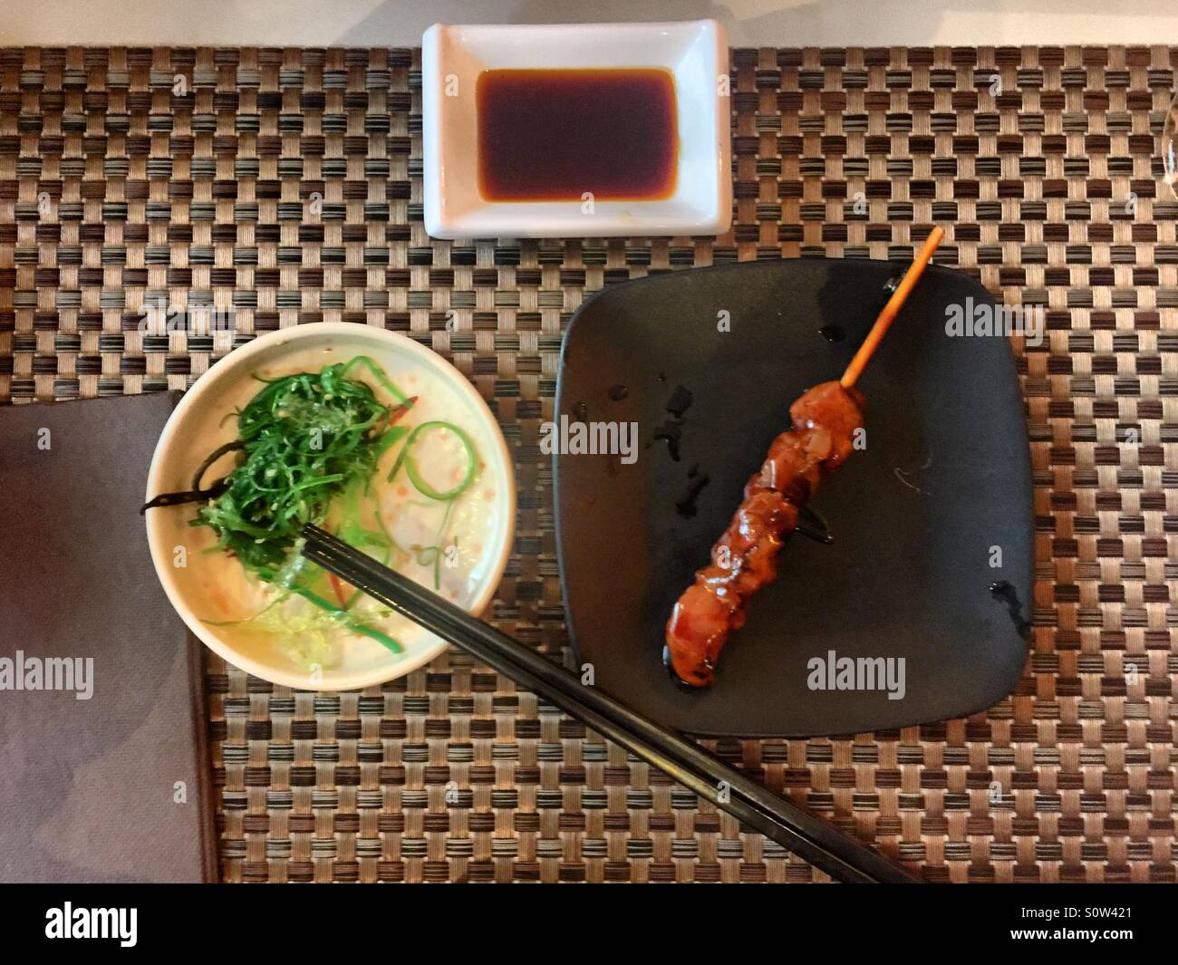 Pasto giapponese con insalata di alghe, uno spiedino yakitori e salsa di soia su un tovaglietta tessuta, flat lay - Immagine stock catturata con smartphone