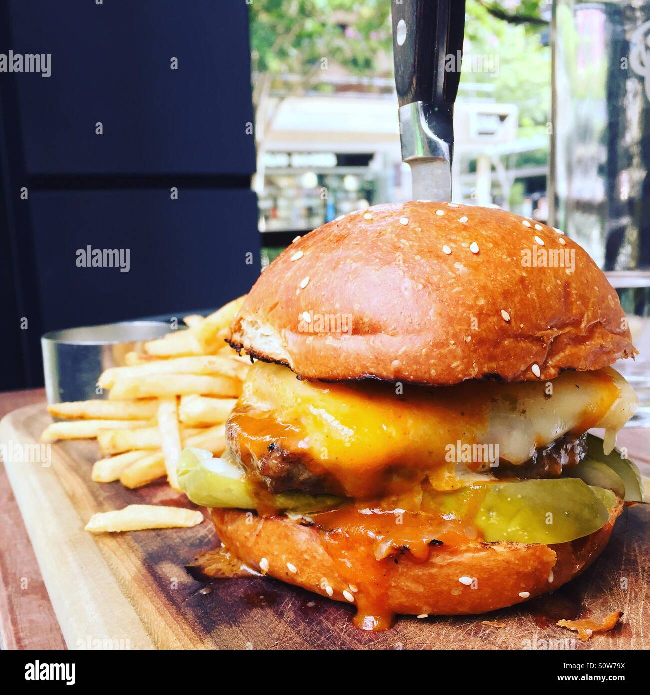 Un gocciolamento di hamburger con formaggio e salsa su una scheda con le patatine fritte o chip, un coltello in esso per mantenerlo. Hamburger sono deliziosi. Foto Stock