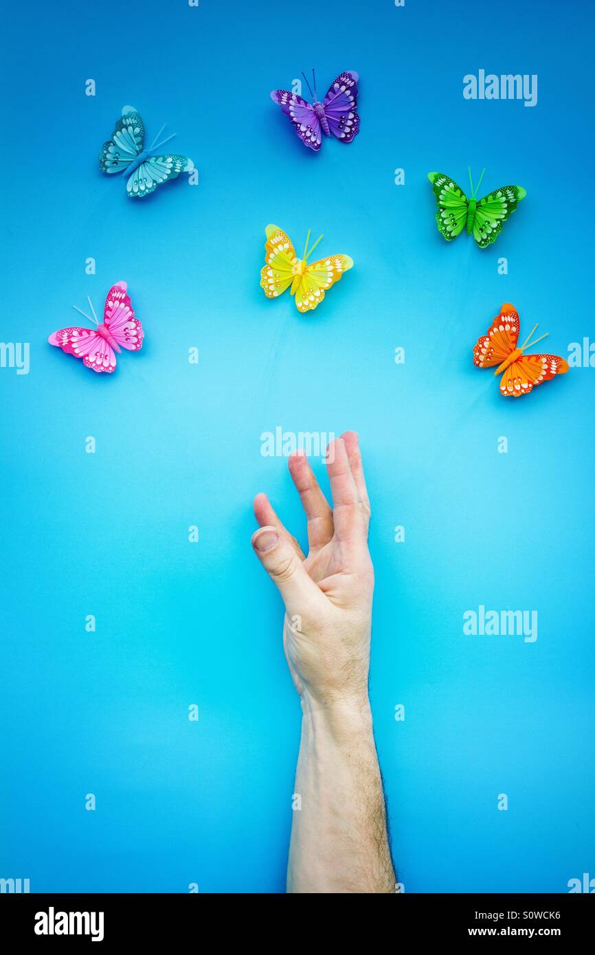 Un uomo la mano e il braccio per raggiungere un gruppo di farfalle variopinte su un luminoso, sfondo blu. Foto Stock