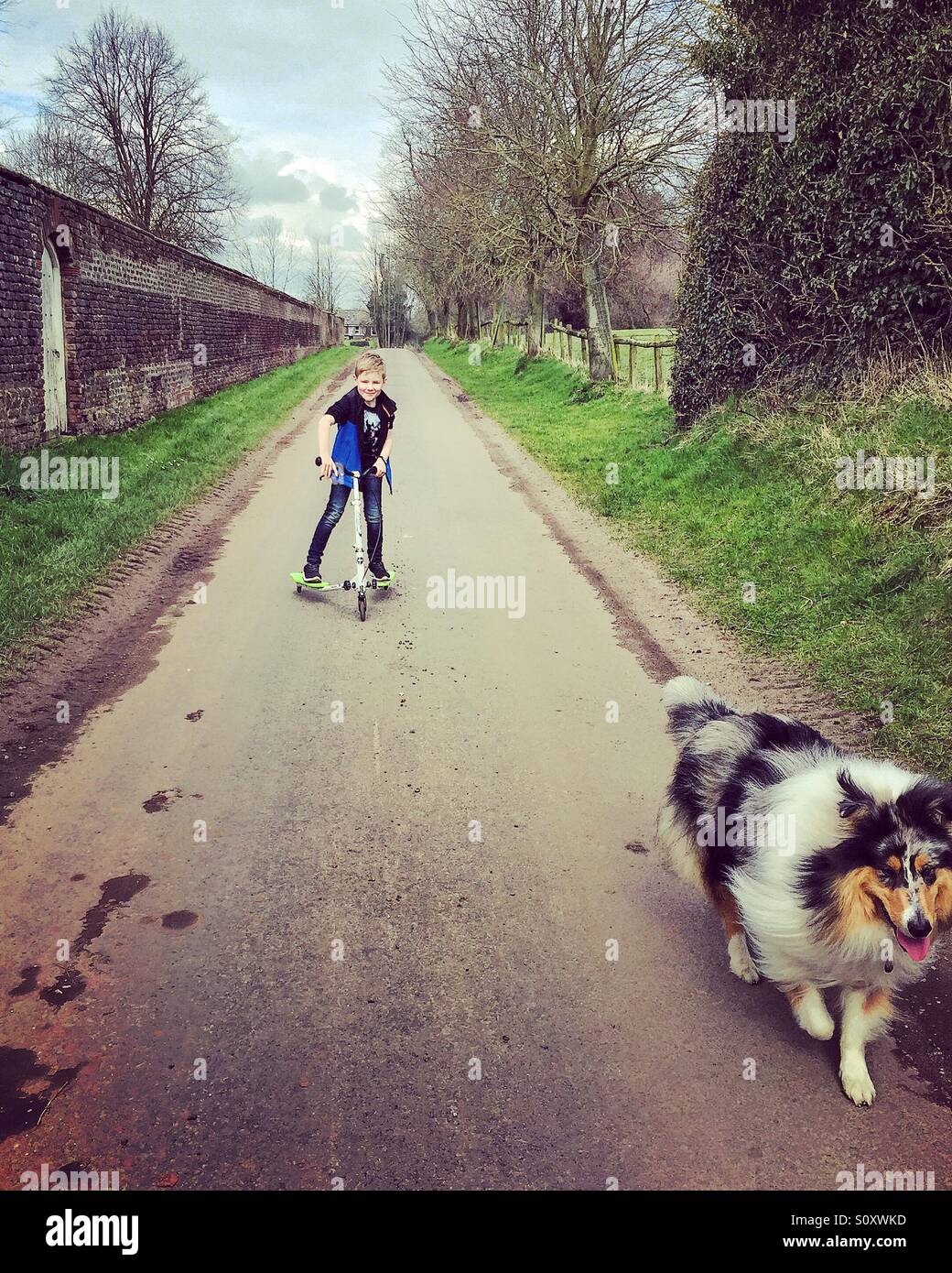 Un ragazzo sul suo scooter seguendo il suo cane lungo una strada di campagna Foto Stock