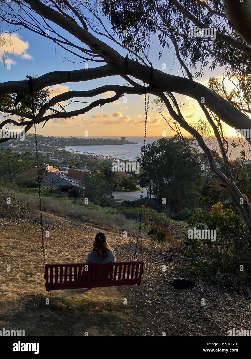 Donna seduta su rustiche panca in legno swing appeso a un albero con una vista perfetta che si affacciano sull'Oceano Pacifico al tramonto. Presa 3o marzo 2016. Foto Stock