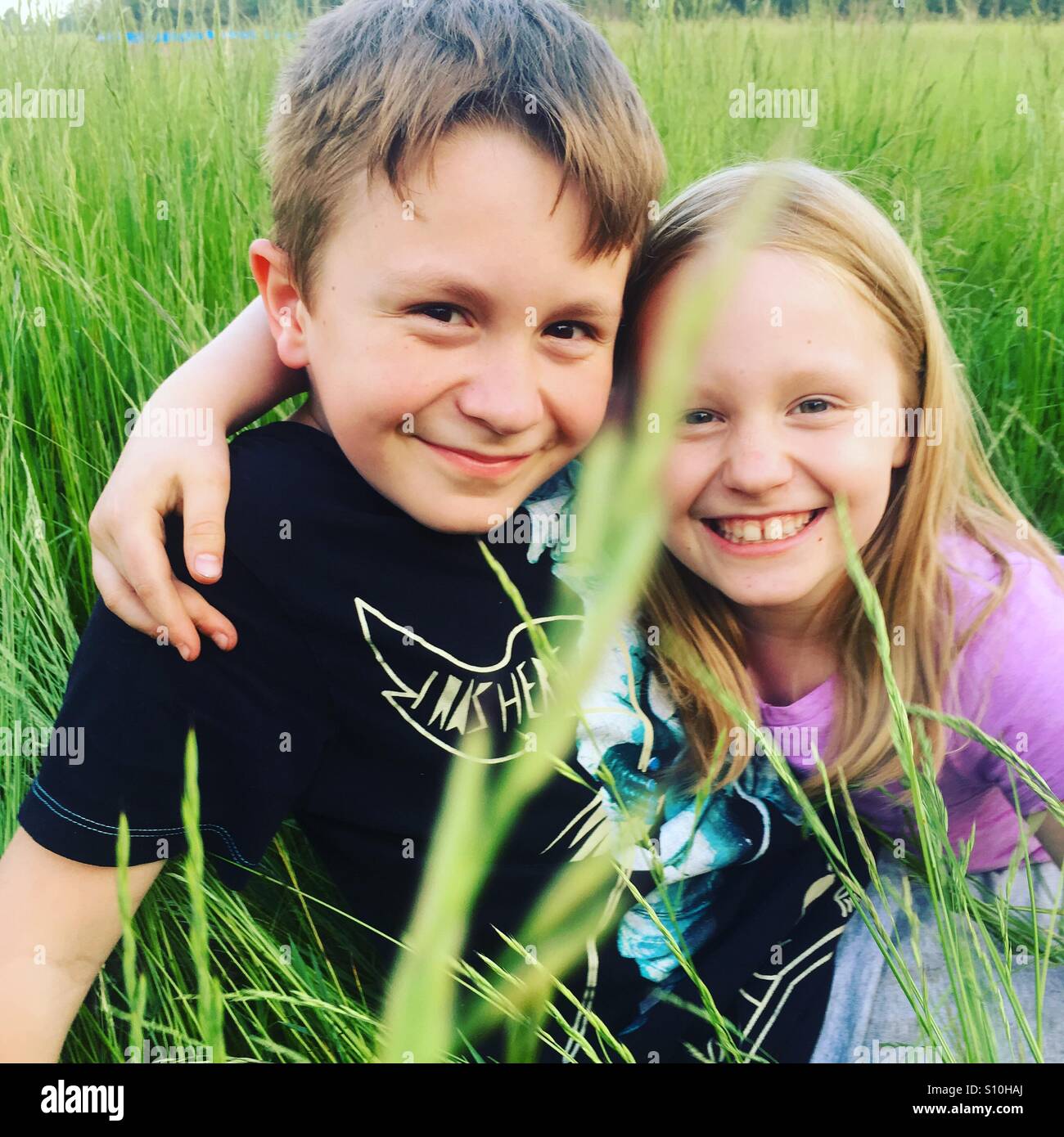 Fratelli di Felice costeggiata a vicenda e sorridente, seduti fuori in un alto erba verde su una multa, bello e soleggiato, fine settimana pomeriggio Foto Stock