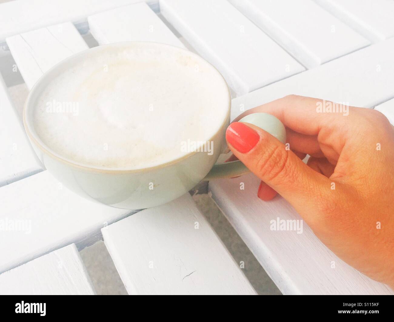 Donna di mano che regge una tazza di cappuccino lattiginoso su un bianco tavolo esterno. Luce naturale. Spazio per la copia. - Immagine stock catturata con smartphone