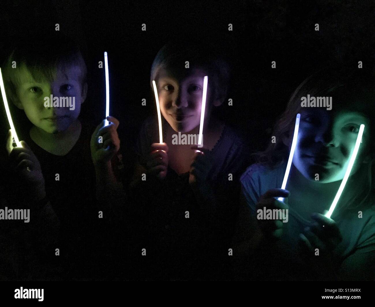 Tre figli illuminare le loro facce con bastoni di incandescenza al buio. Foto Stock