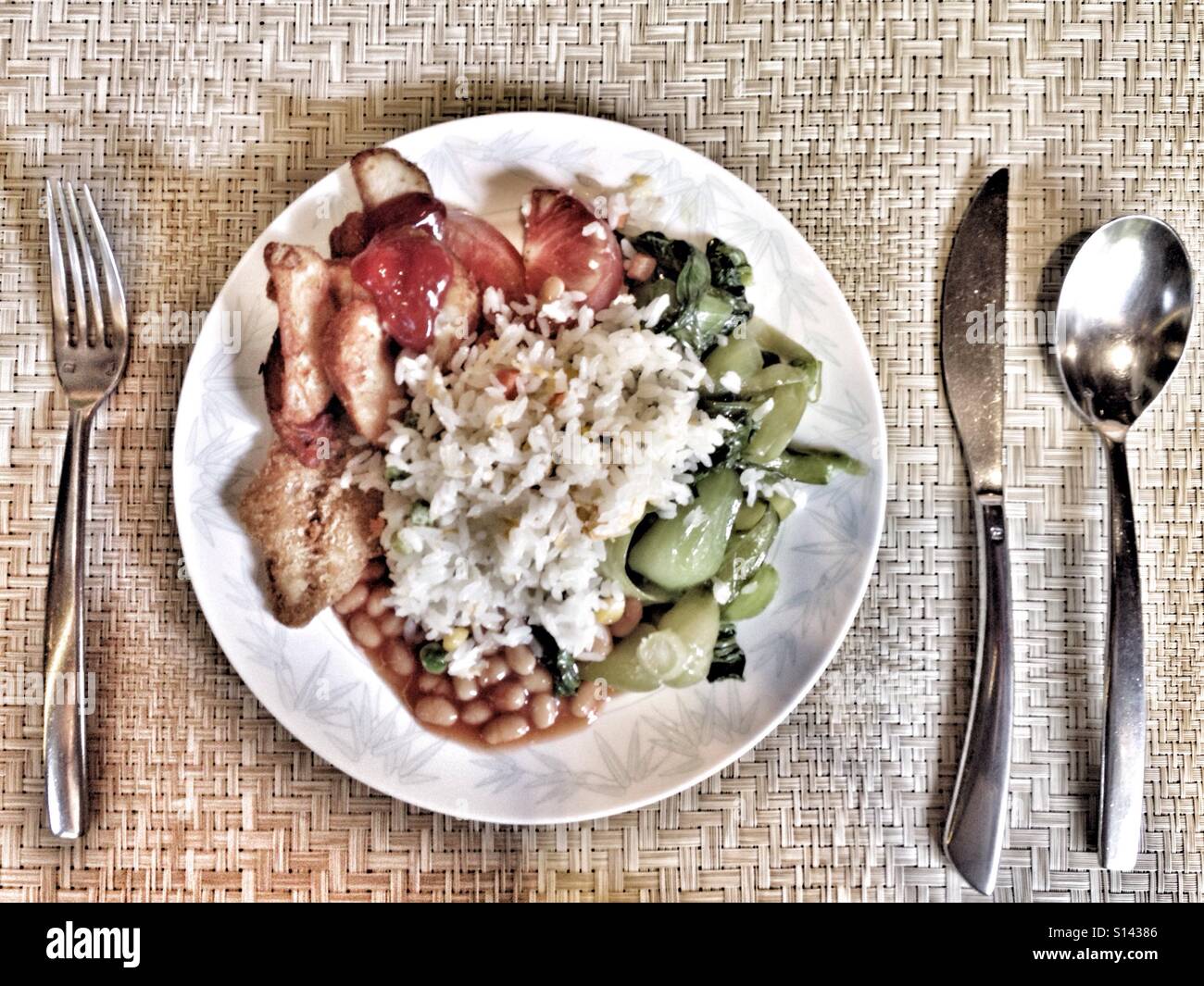 Riso, verdure, patate, fagioli al forno, pomodori sul piatto bianco accanto a cucchiaio, coltello, forchetta sul tappetino di bambù sfondo Foto Stock