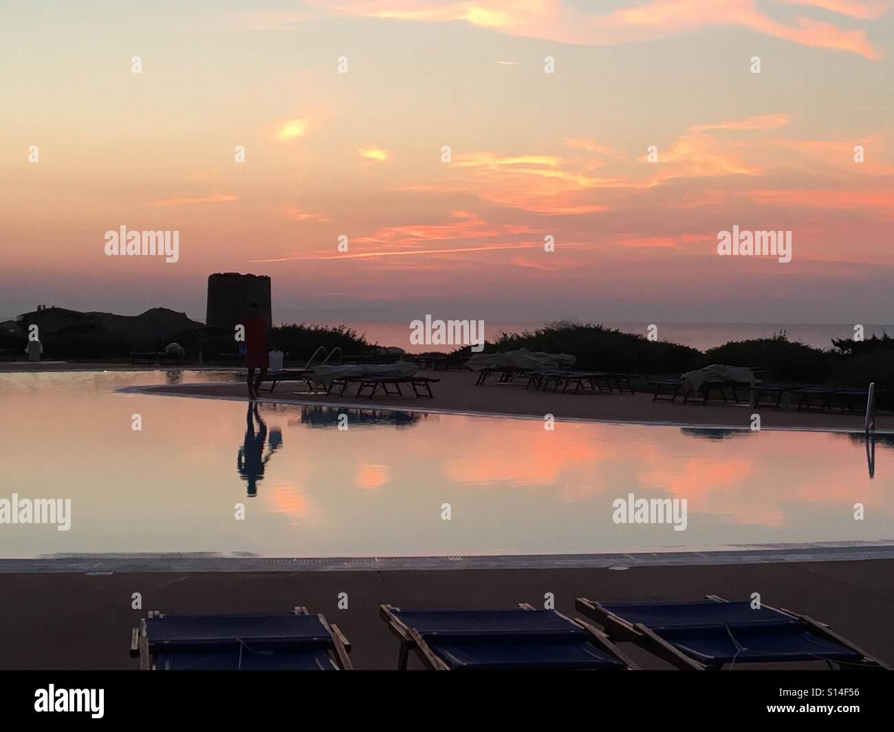 Riflessi di un tramonto colorato oltre l'Hotel Torreruja piscina, l uomo e la torre spagnola sull'Isola Rossa nella parte occidentale della Sardegna Foto Stock