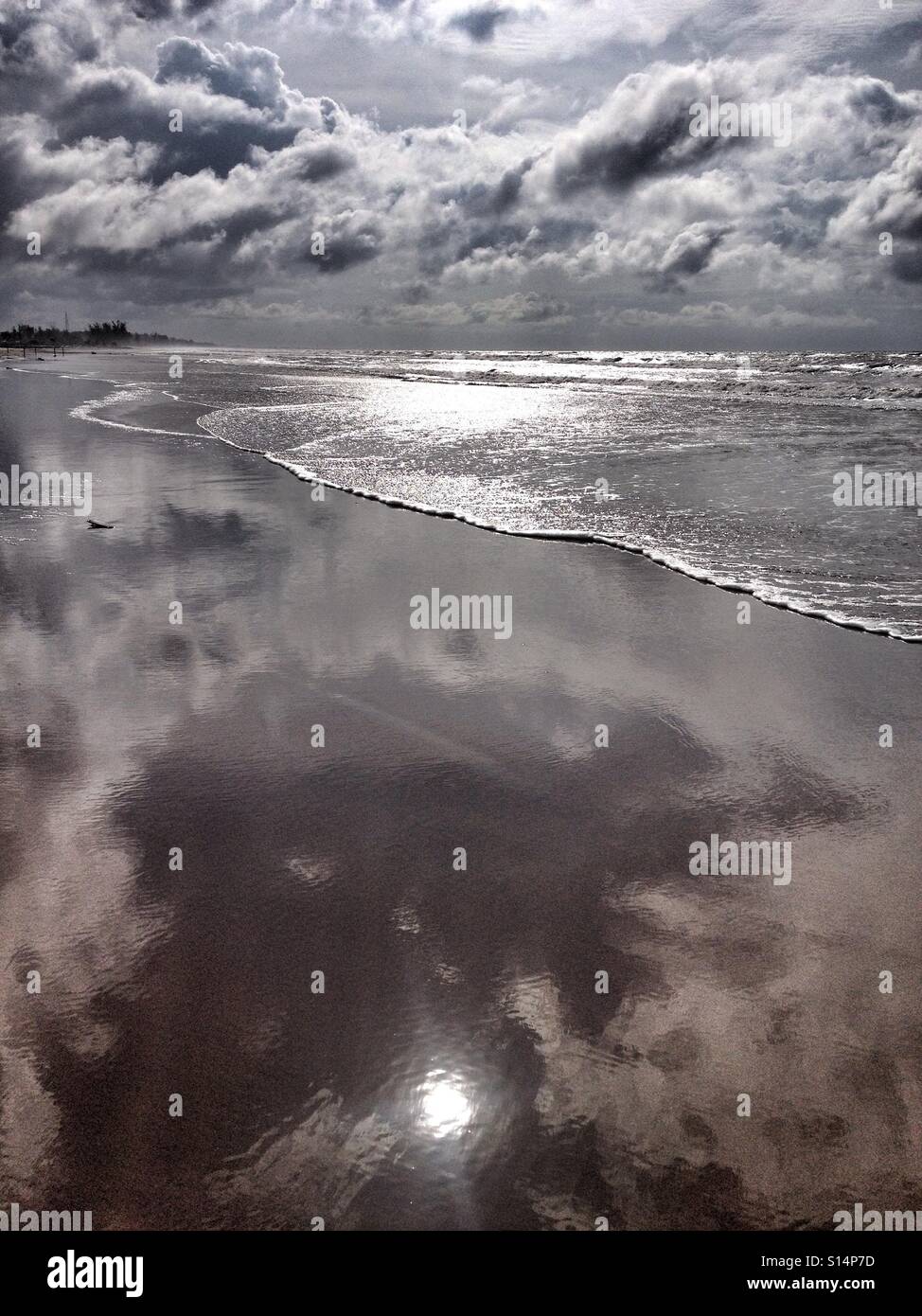 Sabbia bagnata lungo la riva del mare si riflette la luce del sole, come le onde si inclinano indietro nell'oceano, sotto il bianco soffice e cielo molto nuvoloso Foto Stock