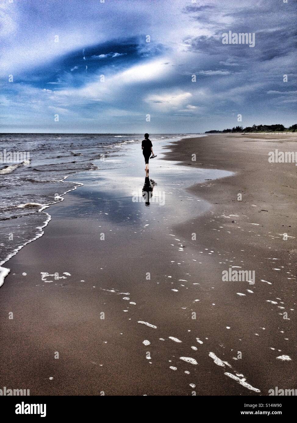 Lone straniero passeggiate lungo le sabbie della spiaggia a piedi nudi, tenendo sandali in mano, come la sabbia bagnata riflette la sua figura Foto Stock