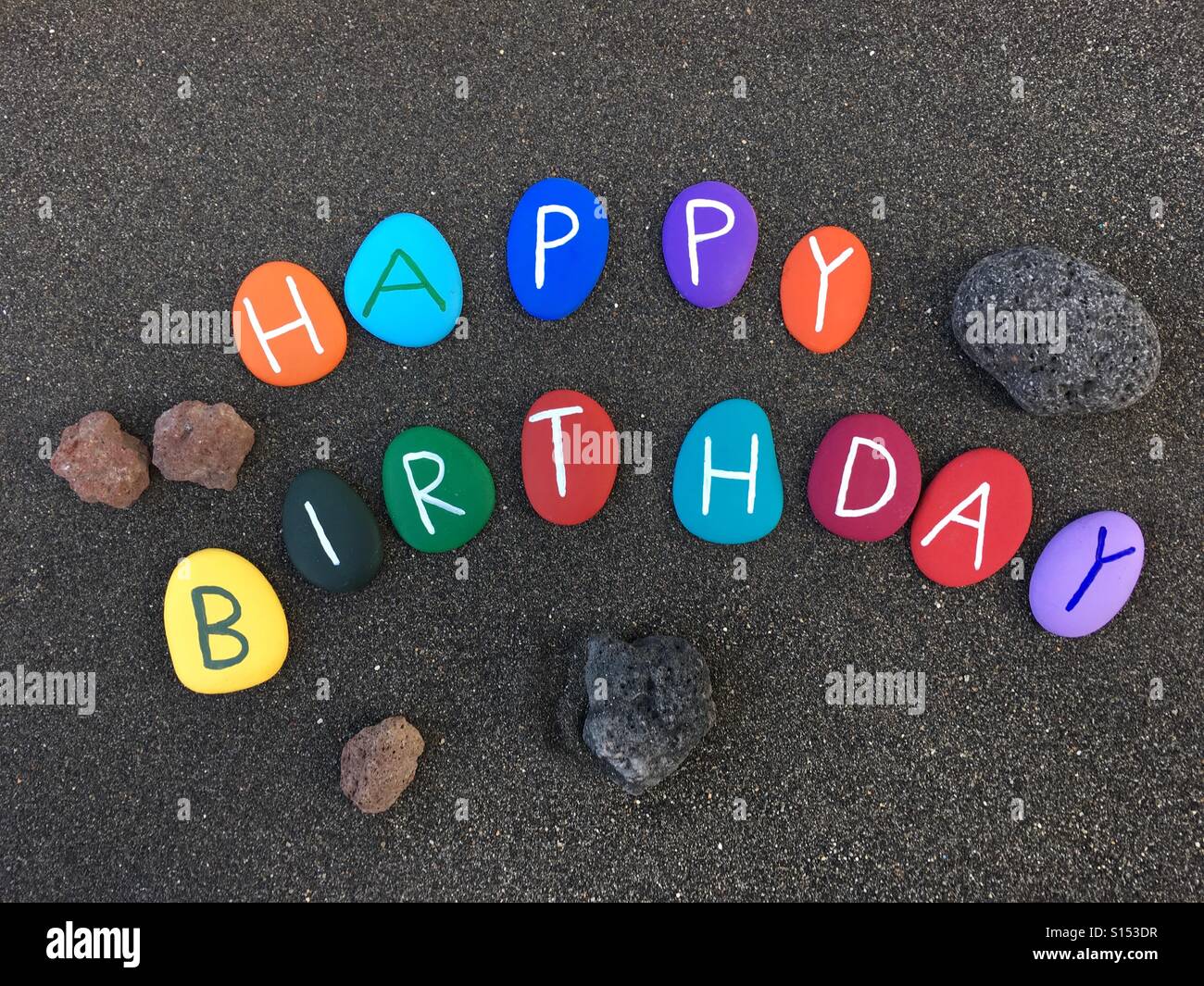 Testo di Happy Birthay composto da lettere in pietra colorata dipinte a mano su sabbia nera naturale dell'isola di Vulcano, isole Eolie, Sicilia, Italia - Immagine stock catturata con smartphone