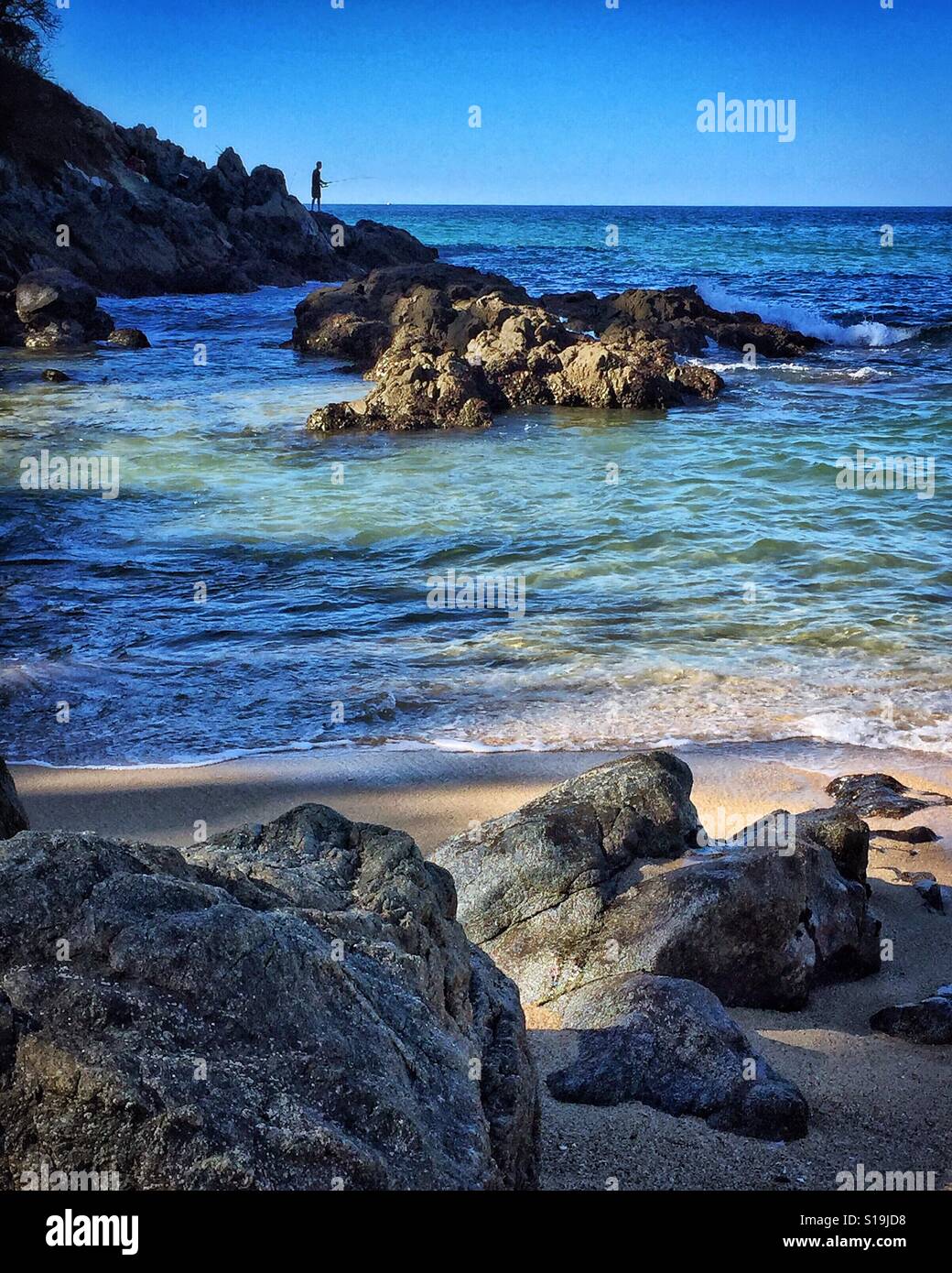 In lontananza un uomo sorge su uno sperone roccioso a punto la pesca su questo robusto e bellissimo tratto di spiaggia della Riviera Nayarit. Foto Stock