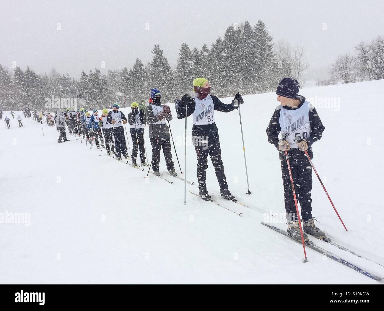 Gli sciatori di fondo si preparano per la partenza di una gara della scuola media durante uno squall di neve. Questi sciatori sono un singolo file su una classica pista di sci nordico. - Immagine stock catturata con smartphone