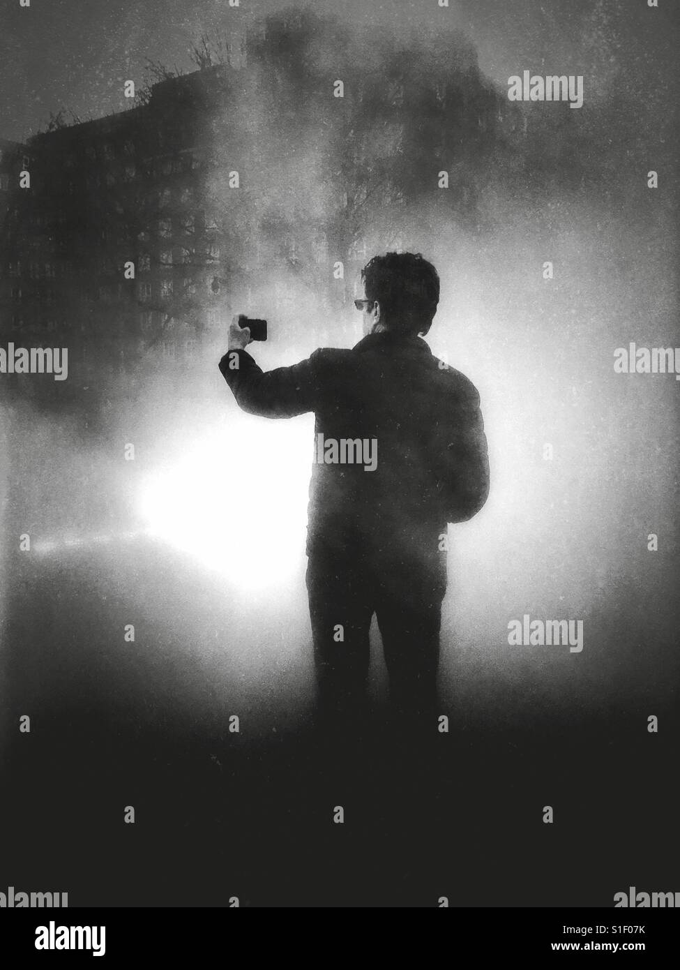 Foto in bianco e nero con illuminazione drammatica di un uomo con il suo telefono cellulare dotato di fotocamera digitale per la lunghezza del braccio mentre posa per una selfie. Foto Stock
