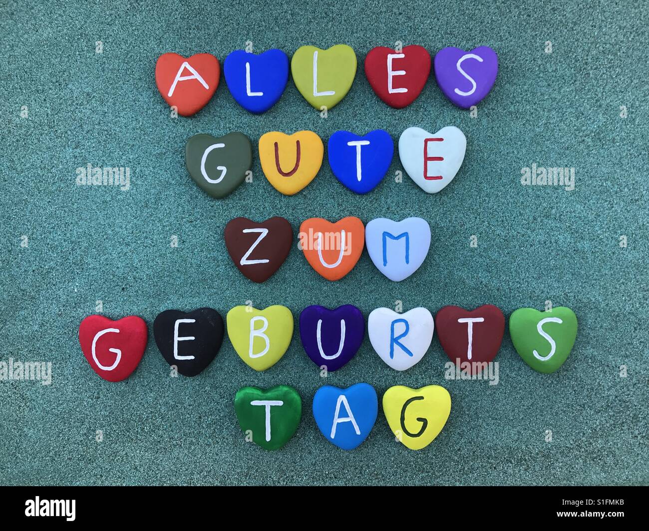 Alles Gute zum Geburtstag, buon compleanno in lingua tedesca con multicolore cuore pietre su sabbia verde Foto Stock