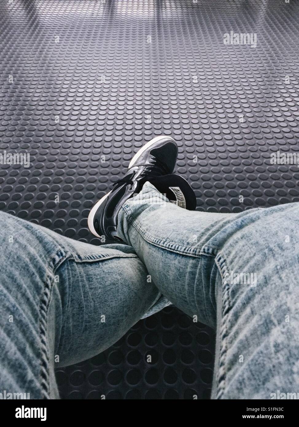 Punto di vista personale, visto attraverso gli occhi di un uomo che guarda i suoi piedi mentre si siede e attende. Indossa un paio di jeans e scarpe da ginnastica. Foto Stock