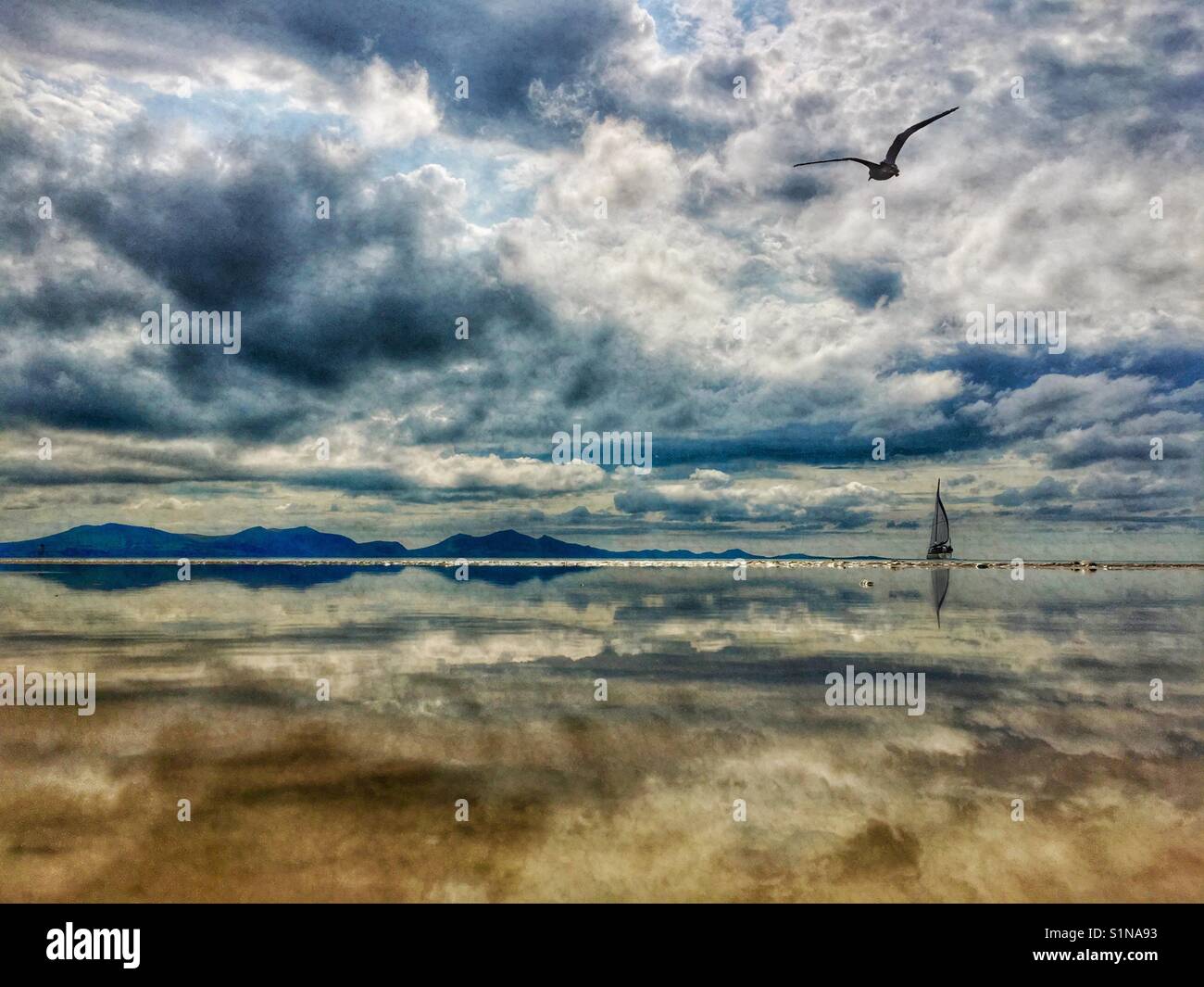 La riflessione di cielo e montagne distanti (Snowdonia) in mare con uno yacht all'orizzonte. Foto Stock