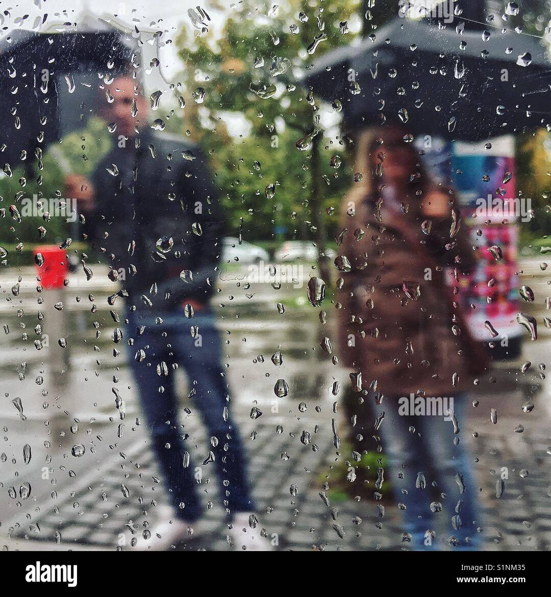 Uomo e donna con ombrelloni attraverso una finestra con le gocce di pioggia - Immagine stock catturata con smartphone