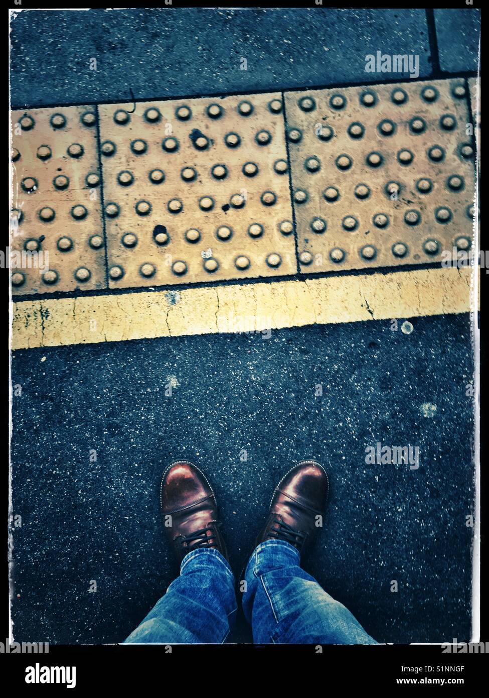 Un maschio di jeans blu e marrone e scarpe in pelle e in piedi su una piattaforma del treno con giallo con pavimentazioni tattili, London, Regno Unito Foto Stock