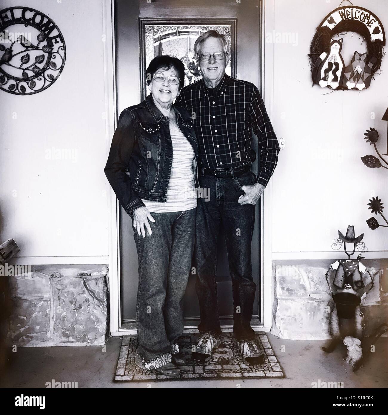 Immagine in bianco e nero di un sorridente caucasian coppia senior in piedi sul tappeto di benvenuto in .davanti a loro la porta anteriore Foto Stock