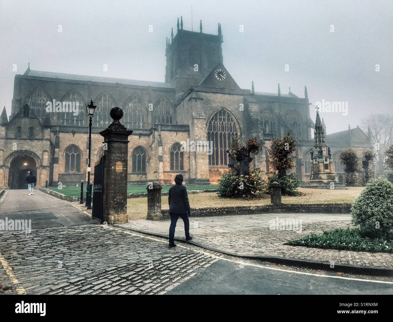 Donna che cammina davanti a Sherborne Abbey, nella nebbia. Sherborne, Dorset, Inghilterra - Immagine stock catturata con smartphone