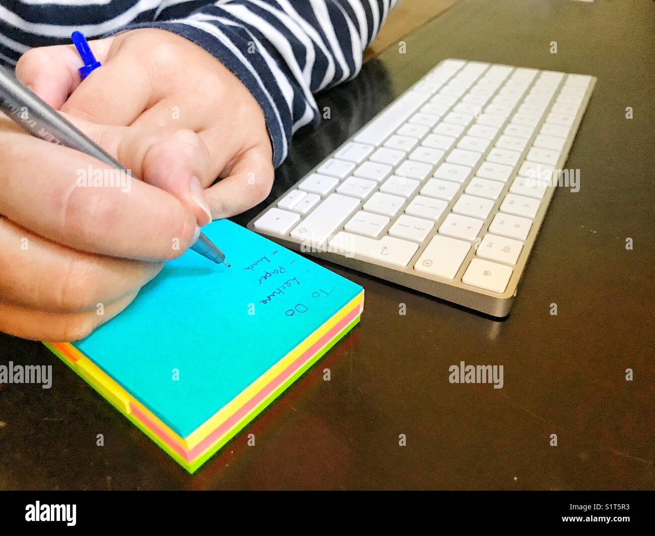 Millennial che scrive un elenco su un blocco appiccicoso, su una scrivania, accanto alla tastiera di un computer - Immagine stock catturata con smartphone