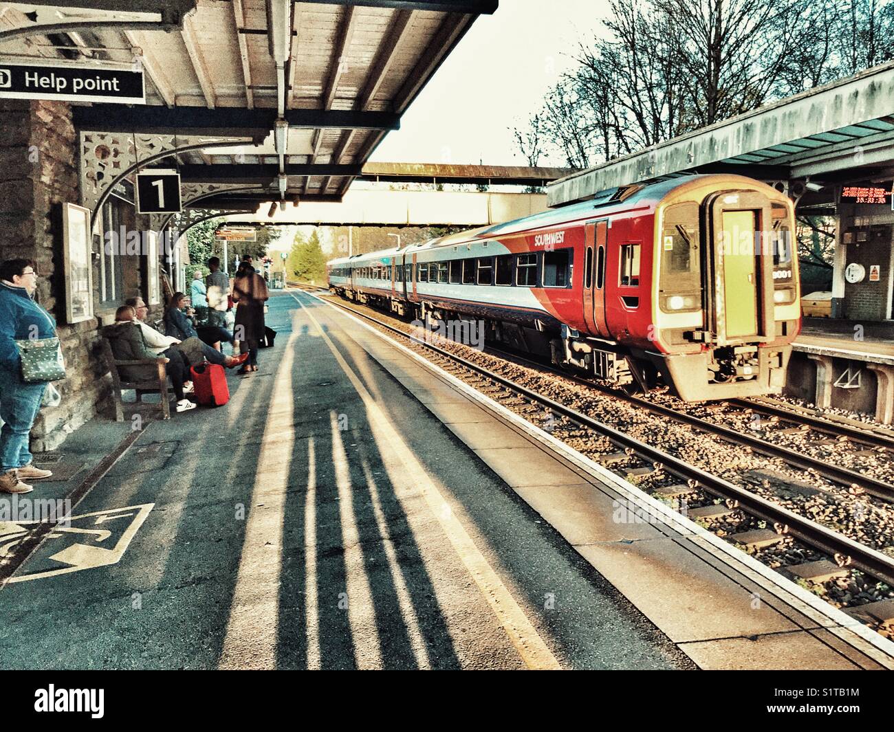 Le persone e le ombre sul rail station piattaforma con un treno in avvicinamento. Sherborne, Dorset, Inghilterra - Immagine stock catturata con smartphone