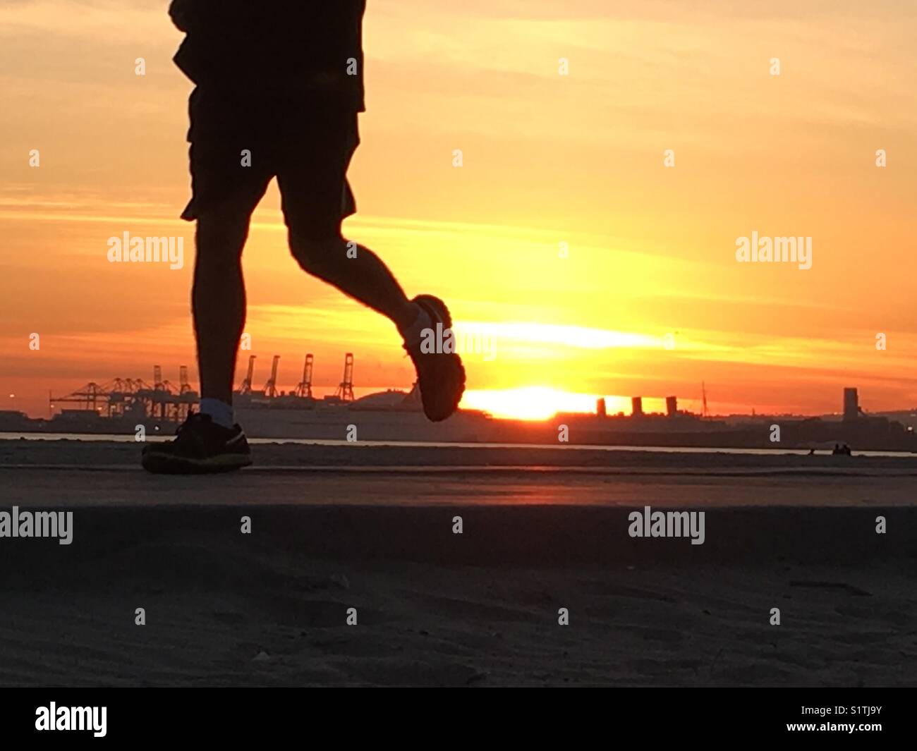 Uomo che corre sulla spiaggia al tramonto con la Regina Maria e porto di Long Beach, CA, Stati Uniti d'America in background. Foto Stock