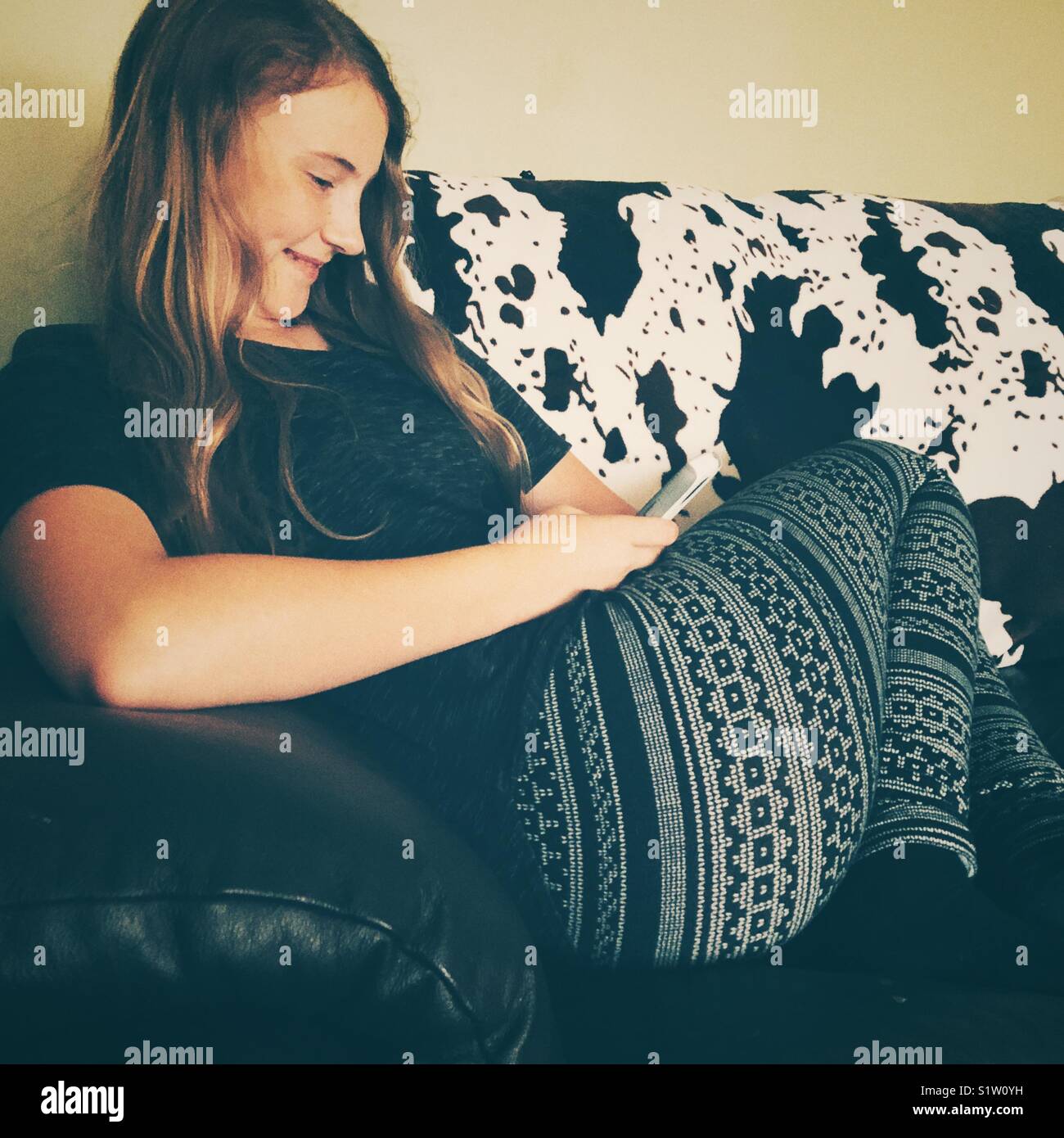 Candida immagine di 12 anni ragazza sorridente a smartphone su un divano con una vacca Holstein coperta di stampa Foto Stock