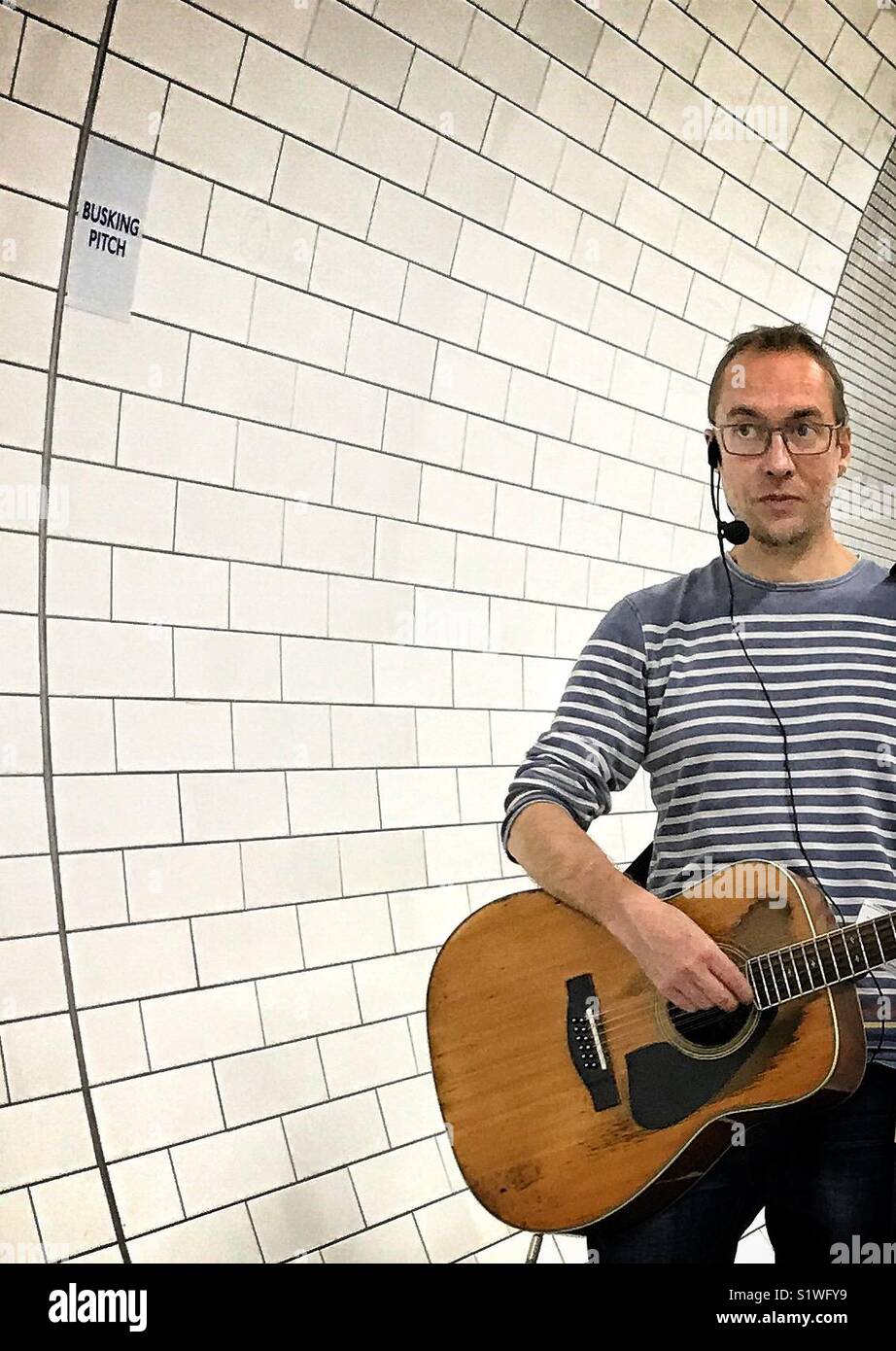 Un suonatore ambulante in un grigio e bianco striato pullover di eseguire con una chitarra acustica in mattonelle bianche nel tunnel della metropolitana di Londra ad un ufficiale di busker pitch Foto Stock