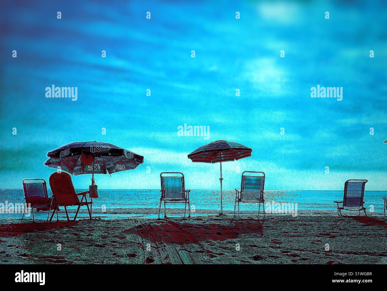 Sedie vuote e ombrelloni su di una spiaggia di sabbia con un mare calmo e azzurro cielo nuvoloso Foto Stock