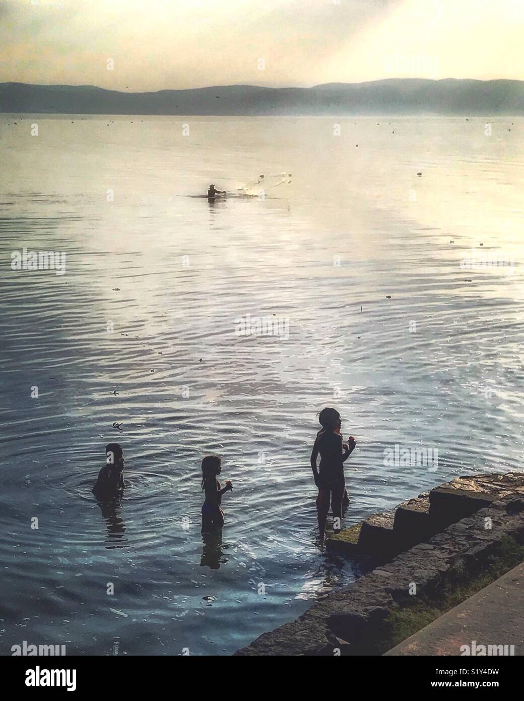 I bambini godono di coni gelato mentre essi wade nel lago in un pomeriggio caldo come un pescatore getta la sua rete per pescare in background. Foto Stock