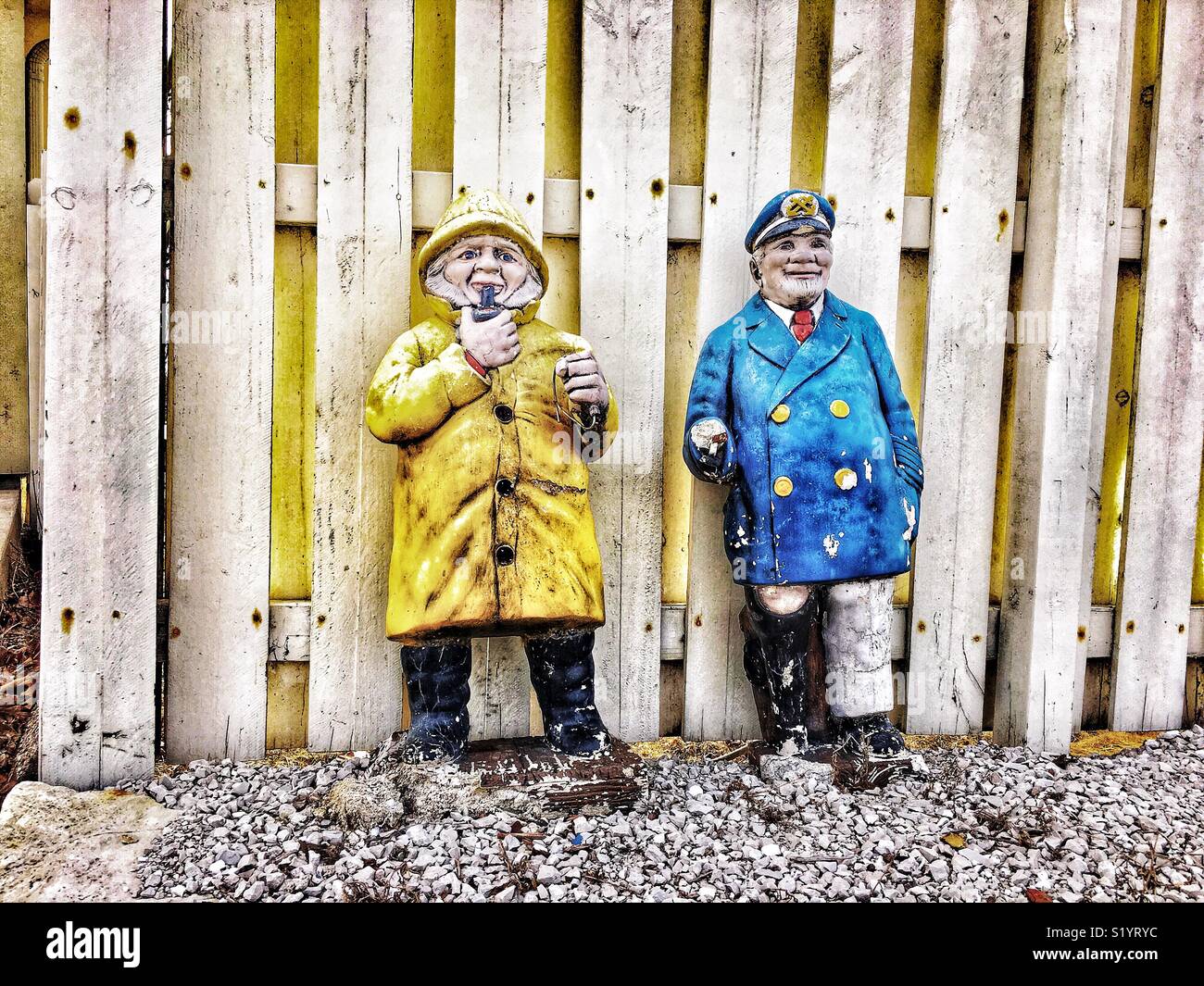 Due ornamenti del prato, una gamba peg marinaio e un uomo fumare in giacca pioggia, appoggiarsi contro Picket Fence sulla strada di ghiaia Foto Stock