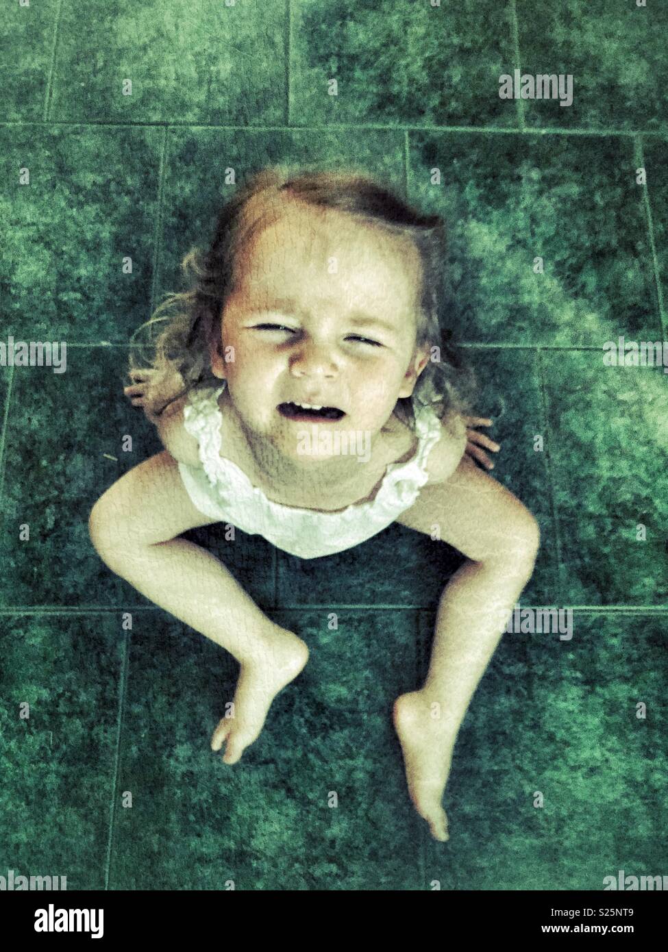 Il Toddler ragazza seduta sul verde piastrelle del pavimento avente una temper tantrum - Immagine stock catturata con smartphone
