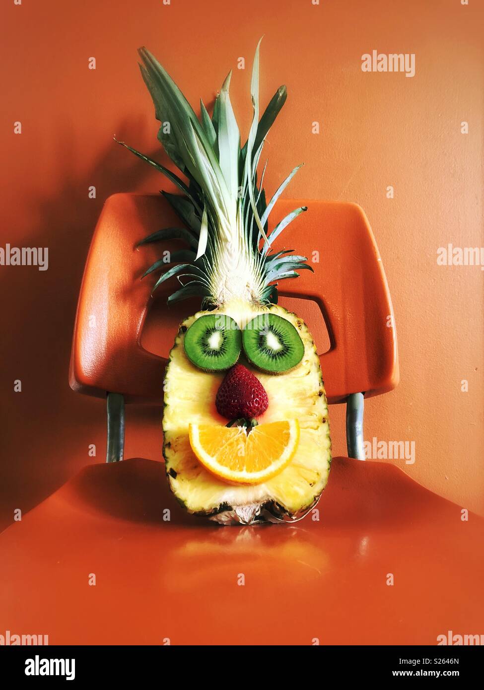 Mezza ananas con fette di kiwi, fragola e fetta di arancia fatto per assomigliare ad una faccia felice seduto su una sedia arancione - Immagine stock catturata con smartphone