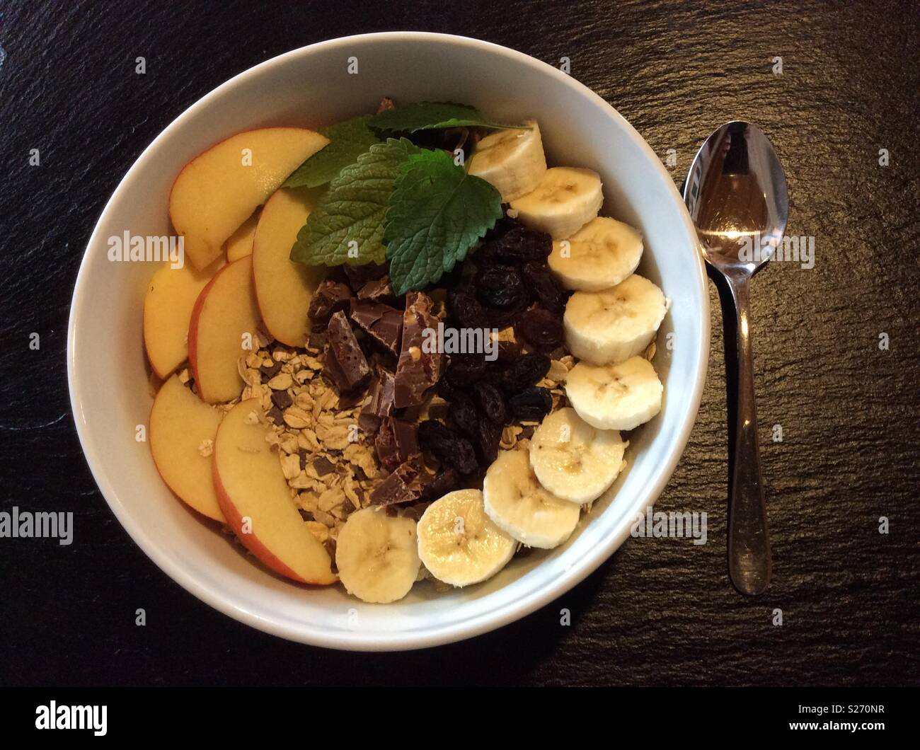 Succulente muesli con banane, cereali, mele, uva passa e cioccolato in tazza bianca per la prima colazione Foto Stock