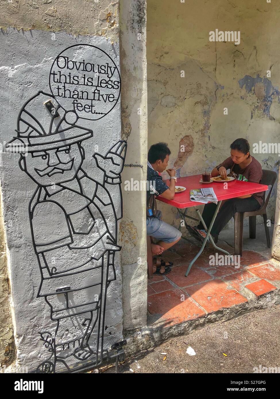 Un locale giovane avente il pranzo accanto a uno di George Town iconici di metallo sculture arte in Penang, Malaysia. Foto Stock
